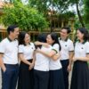 Sáu cựu học sinh tuổi trung niên mặc áo polo trắng phối cổ xanh rêu sọc đỏ ôm nhau mừng rỡ trong sân trường cũ